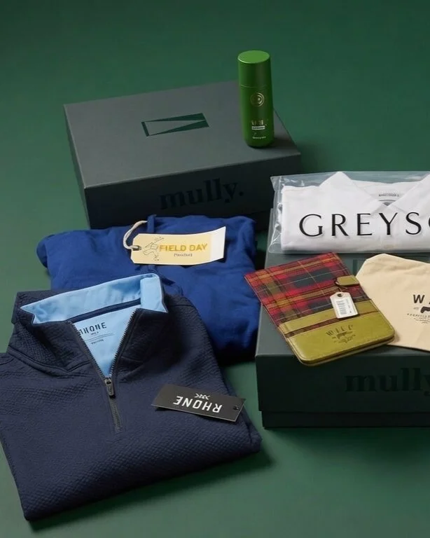 A Mully Reserve quarter laid out on a forest-green editorial surface — Rhone navy quarter-zip, Field Day blue knit, Greyson tee, Will Leather plaid wallet, Penfold canvas bag, and a Will Leather golf shoe carrier.