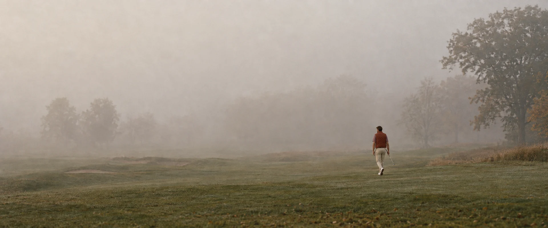 AI-styled editorial scene of a distant golfer wearing a brown quilted vest over a striped polo with tailored khakis on a foggy autumn fairway